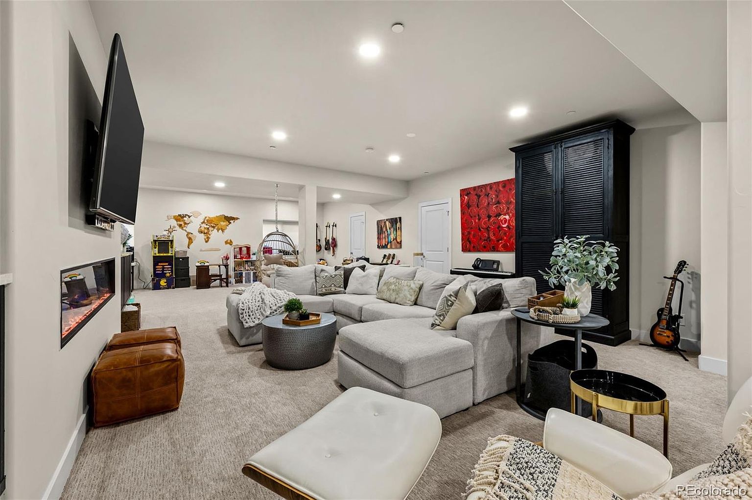 This spacious, finished basement offers a versatile living area featuring a large sectional sofa, a modern fireplace, and a dedicated gaming corner. The room is decorated with vibrant wall art, a world map, and musical instruments, creating a cozy and inviting atmosphere for entertainment. The open-concept layout is well-lit with recessed lighting, making it an ideal space for family gatherings or relaxation.
