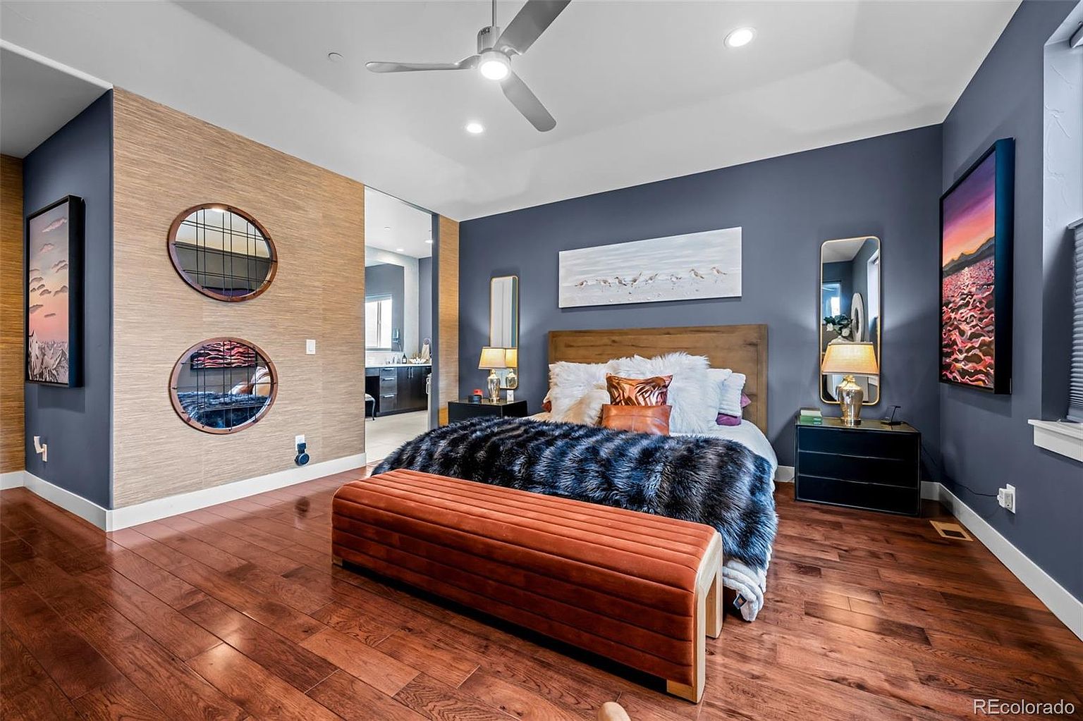 This sophisticated primary bedroom features a striking contrast between deep blue walls and a textured, light-wood accent wall. The room is anchored by a plush bed with a faux-fur throw and a long, velvet-upholstered bench, complemented by modern circular mirrors and bold, colorful artwork. The perspective is a wide, eye-level shot that captures the luxurious, contemporary design and the seamless flow into the adjacent bathroom.