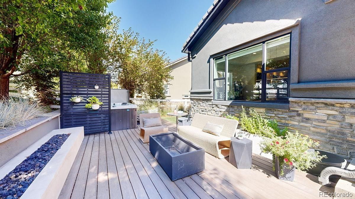 This inviting outdoor living space features a modern composite deck furnished with a stylish wicker sofa, a matching armchair, and a sleek rectangular fire pit. A dark privacy screen separates the seating area from a hot tub, while stone veneer accents on the house exterior add texture and warmth. The scene is bathed in bright, natural sunlight, creating a perfect setting for relaxation and outdoor entertaining.