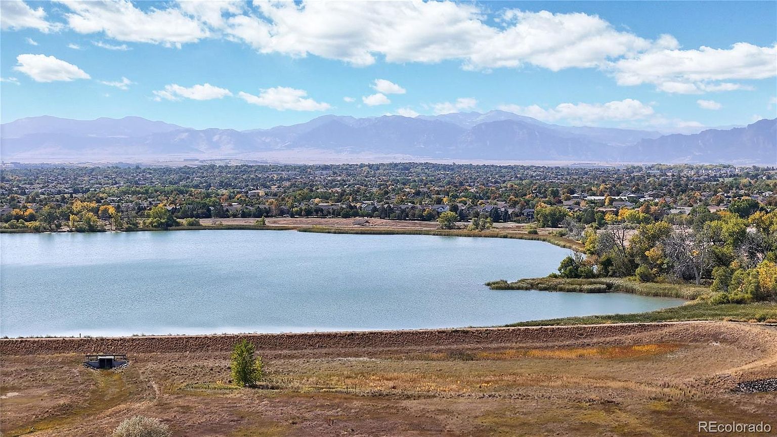 This high-angle aerial view captures a serene lake nestled within a suburban landscape, framed by lush trees and a sprawling residential neighborhood. In the distance, majestic mountain ranges rise against a bright blue sky filled with soft, scattered clouds. The perspective emphasizes the peaceful, scenic location and the expansive natural surroundings of the property.