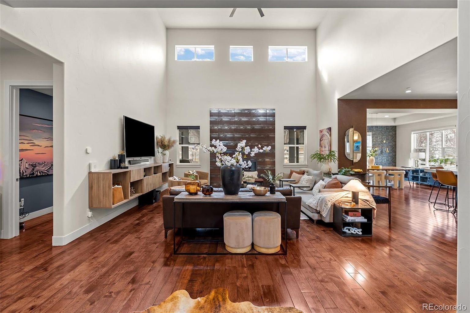 This spacious, high-ceilinged living room features rich hardwood flooring and a striking floor-to-ceiling metal fireplace surround that serves as a dramatic focal point. The open-concept layout seamlessly connects to a dining area, while large windows and clerestory windows flood the space with natural light. The room is tastefully furnished with a contemporary sofa, a console table with ottomans, and a floating media cabinet, creating an inviting and sophisticated atmosphere.