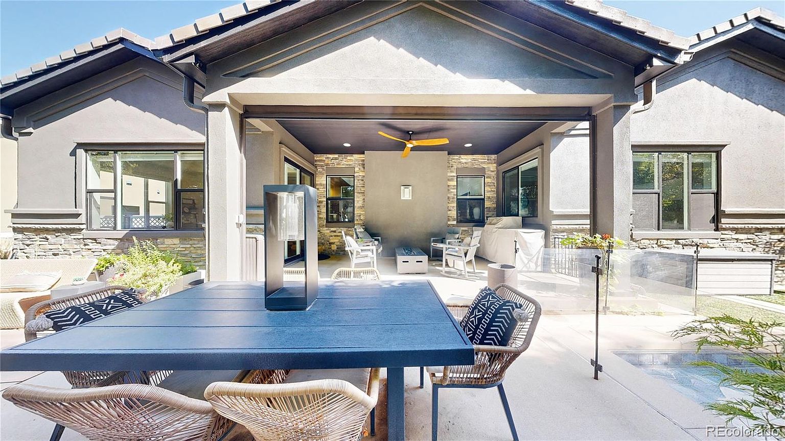 This inviting covered patio features a modern, open-air design with a dark blue ceiling and a central ceiling fan, creating a sophisticated outdoor living space. The area is furnished with a dining table in the foreground and a comfortable seating arrangement near a stone-accented wall, all overlooking a small pool. The perspective is from the dining area looking toward the covered lounge, highlighting a seamless transition between indoor and outdoor living.