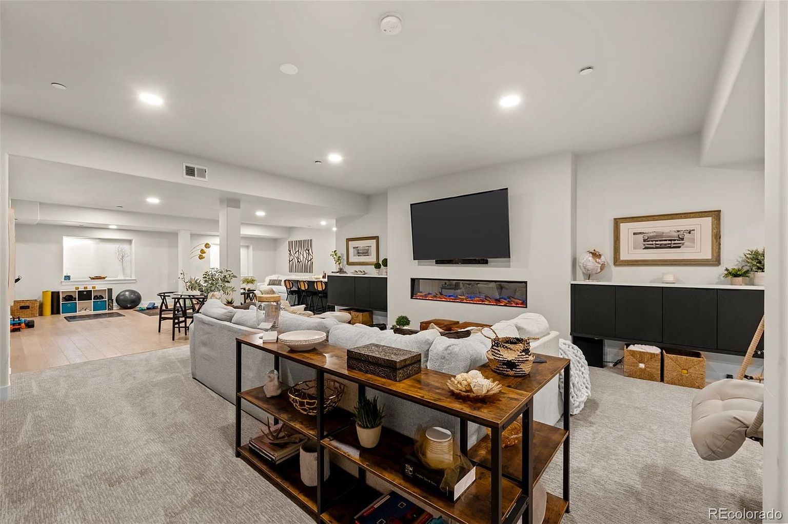 This spacious, open-concept basement features a cozy living area with a large sectional sofa, a modern linear fireplace, and a wall-mounted television. The space transitions seamlessly into a multi-purpose area with light wood flooring, a dining set, and a dedicated workout corner, creating a versatile and inviting entertainment zone. The neutral color palette and recessed lighting contribute to a bright, contemporary, and comfortable atmosphere.