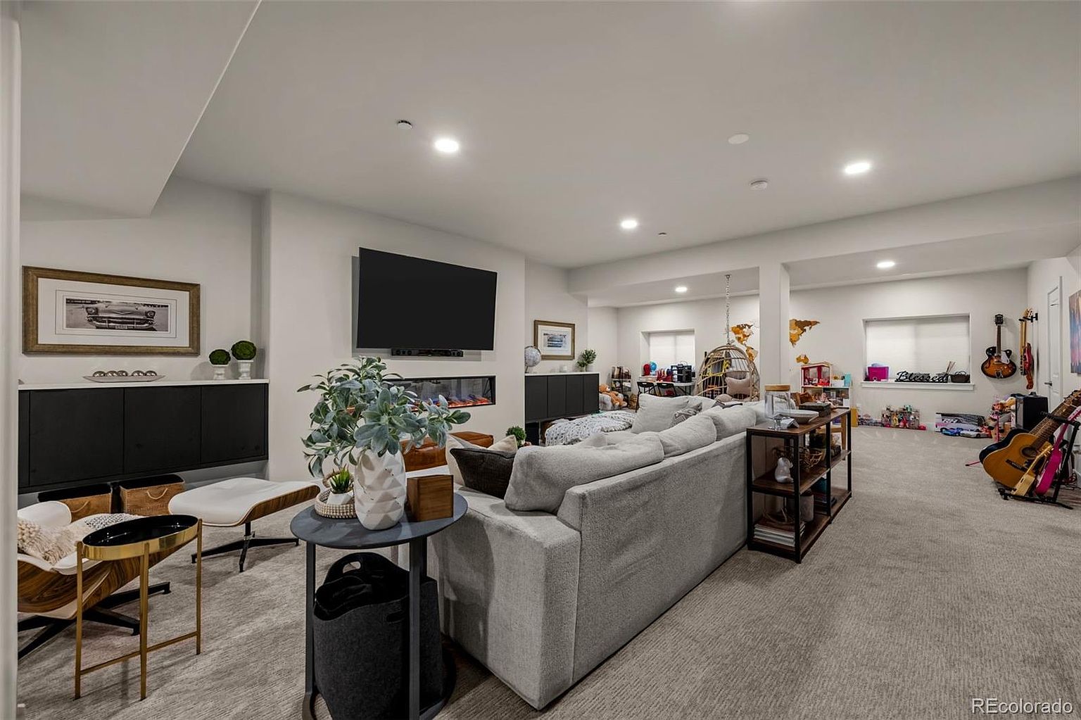 This spacious, finished basement features a comfortable living area with a large sectional sofa, a modern fireplace, and a wall-mounted television. The open-concept layout includes a dedicated play area, a hanging chair, and a corner for musical instruments, creating a versatile and inviting family space. The neutral color palette and recessed lighting provide a bright, contemporary feel throughout the room.