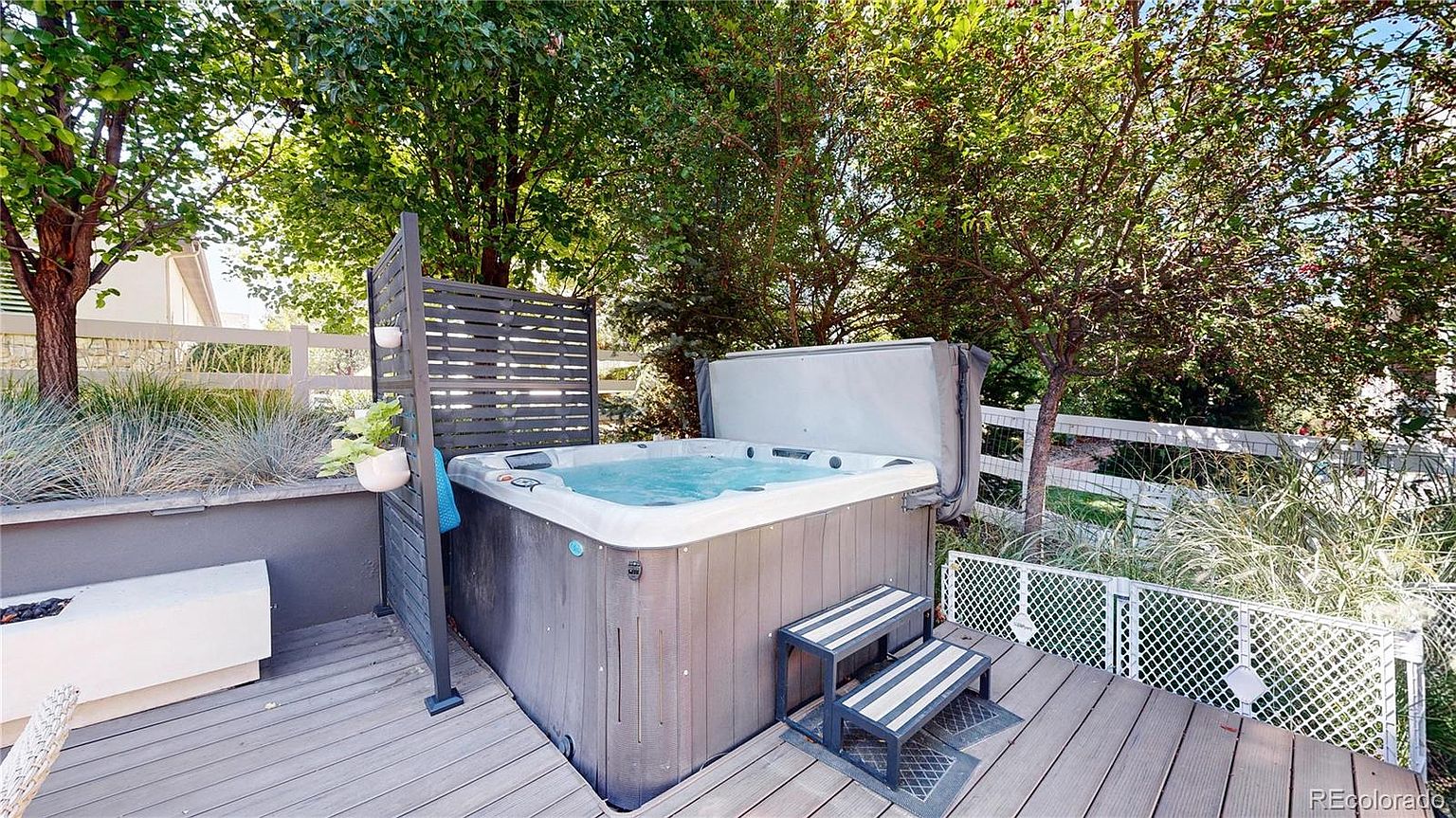 This outdoor space features a modern hot tub set on a spacious wooden deck, providing a private and relaxing retreat. The area is surrounded by lush green trees and a white fence, with a privacy screen attached to the side of the spa for added seclusion. A set of steps leads up to the hot tub, creating an inviting atmosphere for leisure and entertainment.