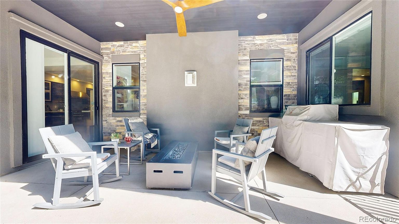 This inviting covered patio features a modern aesthetic with stone accent walls, a sleek fire pit, and comfortable rocking chairs. The space is well-lit with recessed lighting and a ceiling fan, creating a perfect outdoor living area for relaxation or entertaining. A large grill covered in a protective tarp sits to the side, highlighting the functional design of this outdoor retreat.