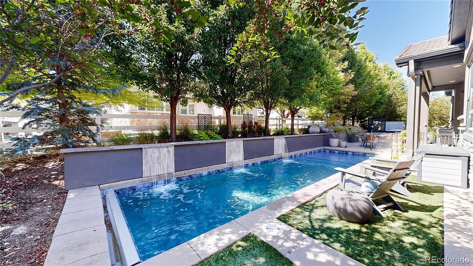 This inviting backyard features a sleek, rectangular lap pool accented by three modern water features cascading from a raised stone wall. The pool area is complemented by a small patch of artificial turf, comfortable lounge seating, and a covered patio space, all framed by lush green trees and a white fence. The scene captures a serene, resort-like atmosphere perfect for outdoor relaxation and entertaining.
