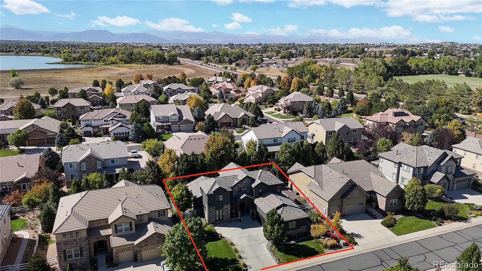 This high-angle aerial view captures a suburban residential neighborhood featuring a prominent two-story home highlighted by a red outline. The property showcases a multi-gabled roofline, a stone and dark siding facade, and a spacious driveway, all set within a well-manicured community with mature trees and a nearby lake. The expansive perspective emphasizes the home's location within the quiet, scenic development against a backdrop of distant mountains.