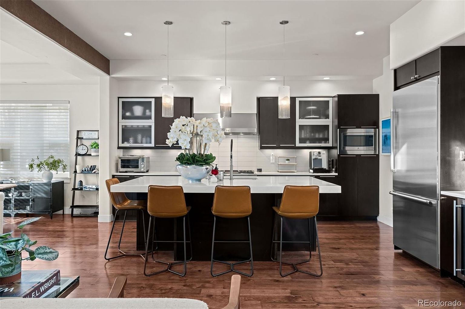 This modern, open-concept kitchen features dark cabinetry contrasted by a large white quartz island with seating for four. The space is illuminated by three sleek pendant lights and recessed lighting, highlighting the warm hardwood floors and stainless steel appliances. A decorative shelf unit and a large floral centerpiece add a touch of personality to this clean, contemporary culinary space.
