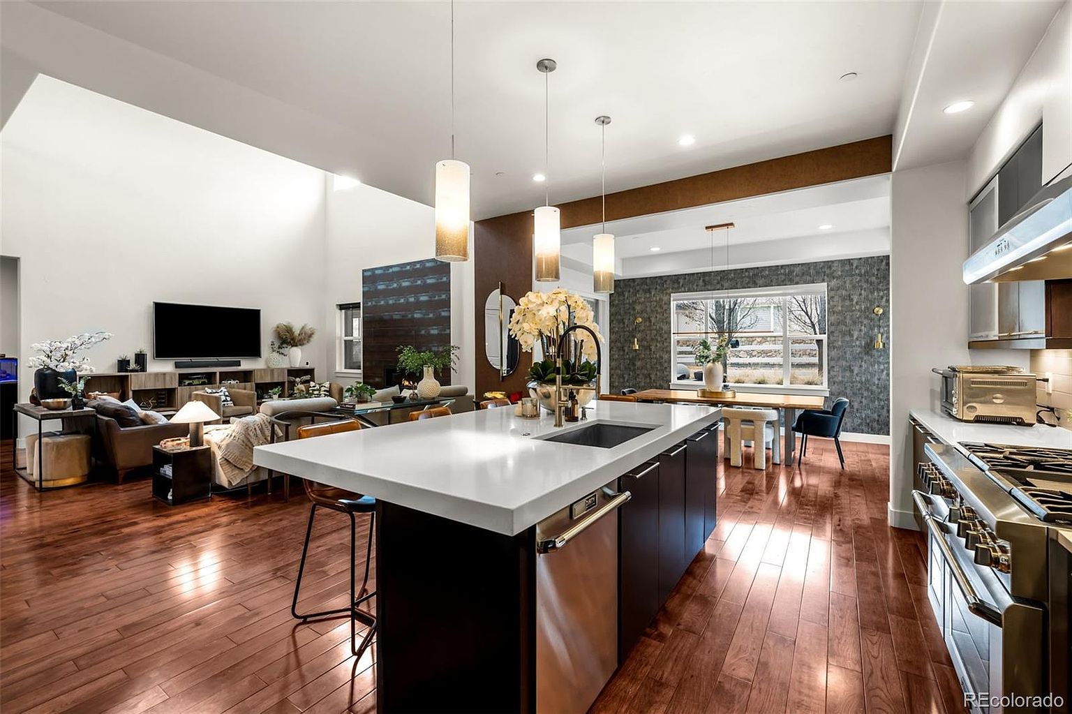 This spacious, open-concept kitchen features a large central island with a white quartz countertop and a stainless steel dishwasher, serving as the focal point of the room. The space is illuminated by three modern pendant lights and flows seamlessly into a bright living area with high ceilings and a dining space in the background. Rich hardwood flooring, dark cabinetry, and high-end stainless steel appliances create a sophisticated, contemporary aesthetic perfect for entertaining.