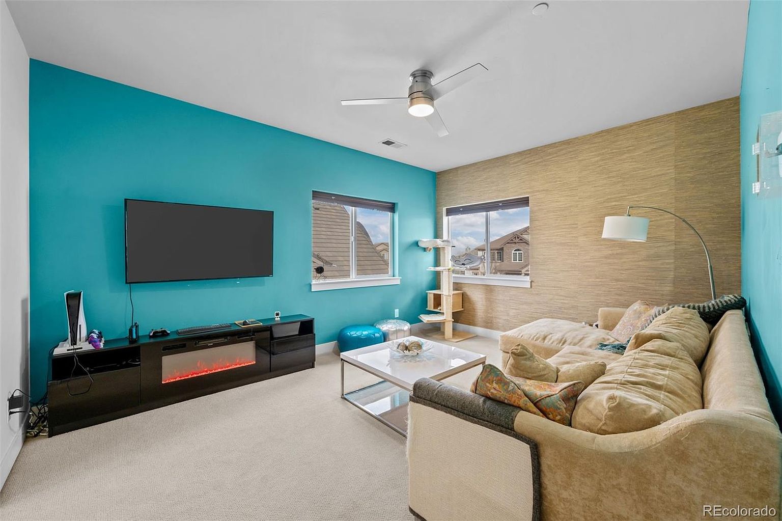 This vibrant living room features a bold teal accent wall contrasted by a textured, neutral-toned wall, creating a modern and inviting atmosphere. The space is furnished with a plush, oversized sectional sofa, a sleek glass-top coffee table, and a media console with an integrated electric fireplace. Large windows allow for ample natural light, highlighting the clean carpeted floors and the contemporary ceiling fan.