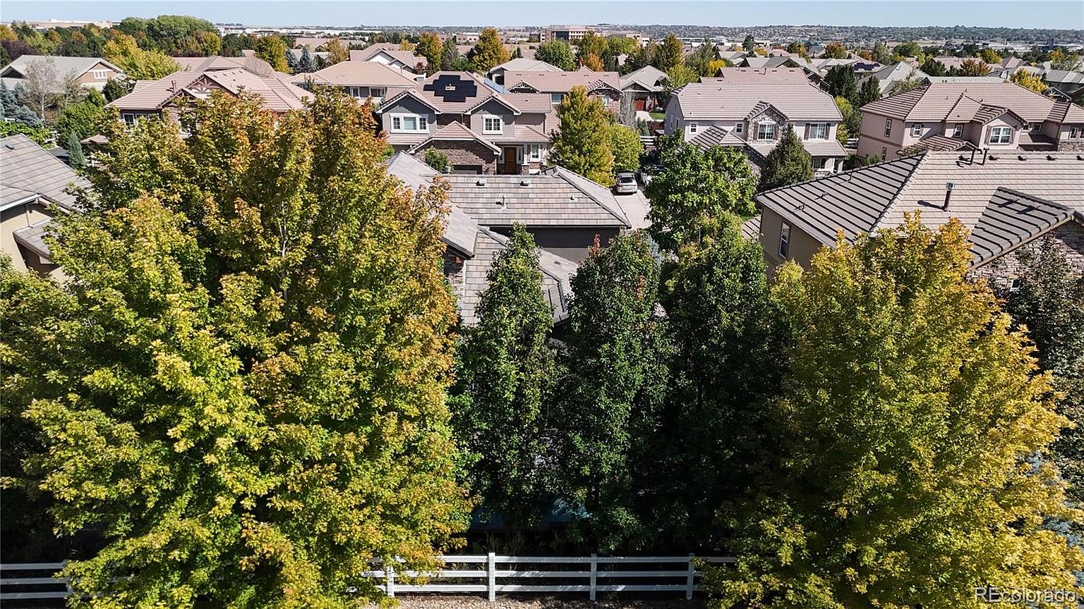 This high-angle aerial view captures a suburban neighborhood characterized by large, multi-story single-family homes with tiled roofs. The foreground is dominated by lush, mature deciduous trees that provide privacy, while a white split-rail fence marks the property boundary. The scene conveys a peaceful, established residential atmosphere under a clear blue sky.