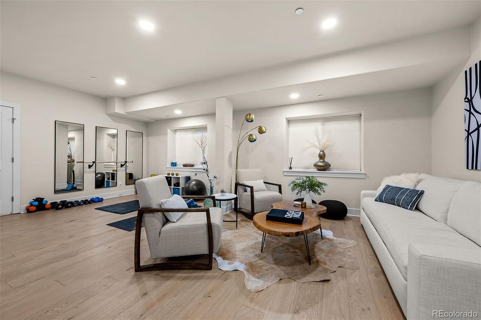This versatile basement space features a bright, open-concept layout that functions as both a cozy living area and a home gym. The room is anchored by a light-colored sofa, two armchairs, and a natural wood coffee table, all set upon a cowhide rug, while the background includes a dedicated workout area with mirrors and a ballet barre. The neutral color palette, recessed lighting, and light wood-look flooring create a clean, inviting, and multi-functional atmosphere.