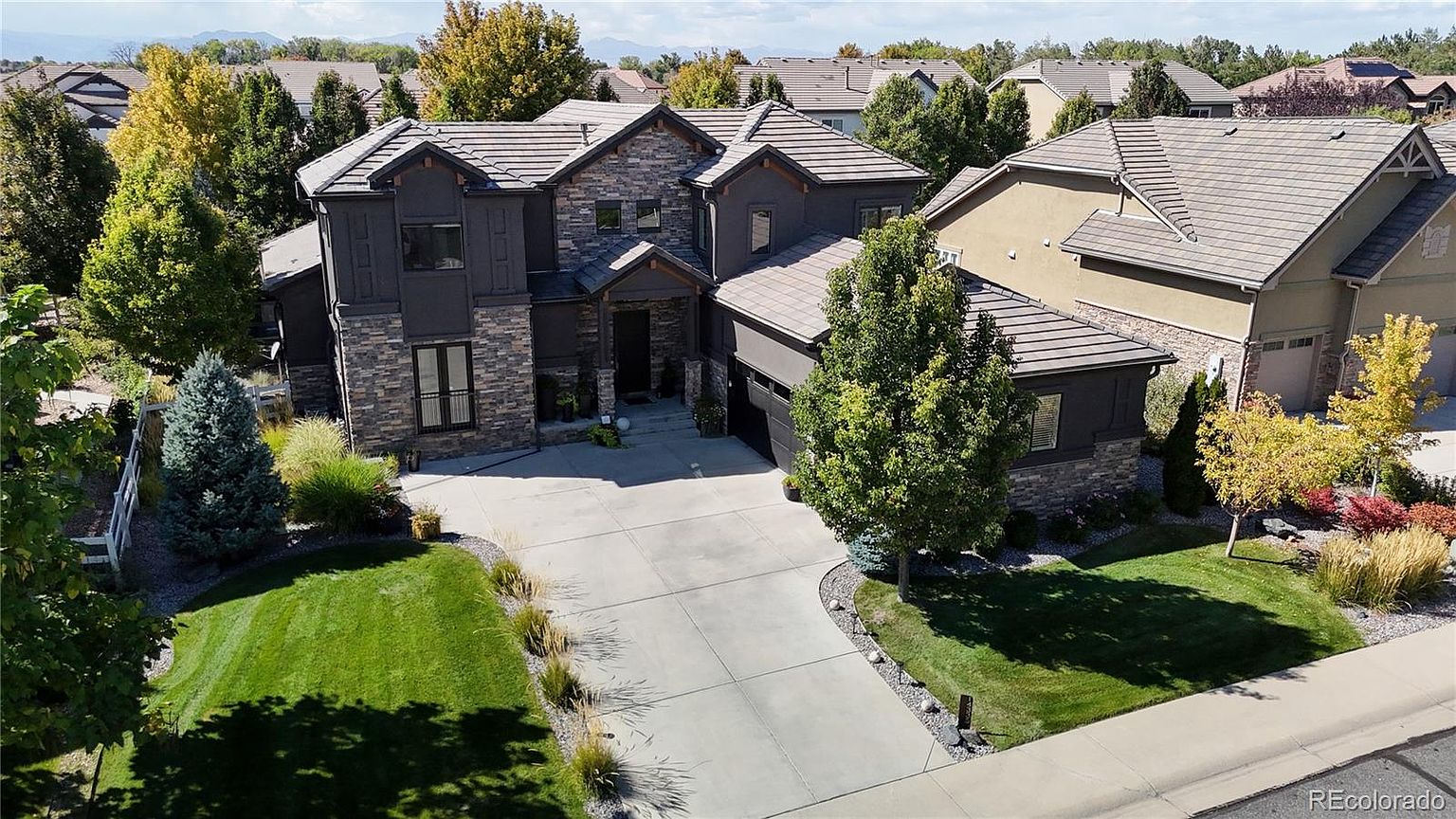 This high-angle aerial shot captures a stately two-story suburban home featuring a sophisticated blend of stone veneer and dark-toned siding. The property includes a spacious concrete driveway, a well-manicured front lawn with mature landscaping, and a multi-car garage. The architectural design emphasizes a grand entryway and a complex roofline, presenting an inviting and upscale curb appeal.