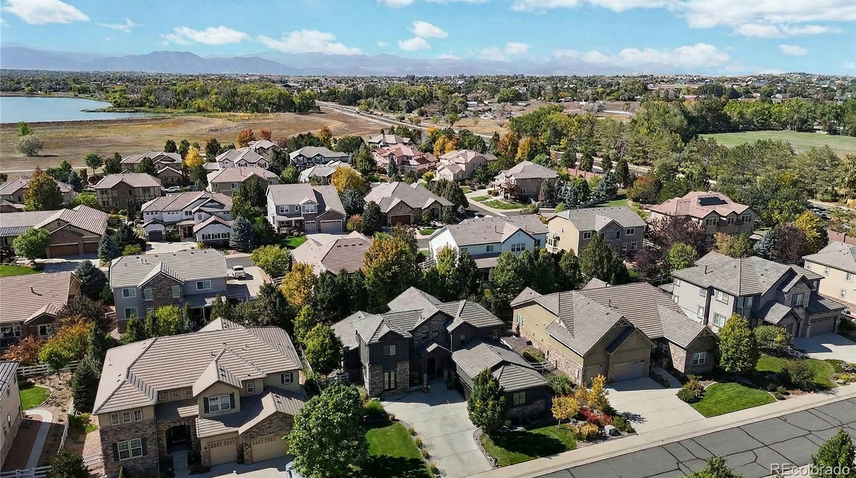 This aerial view captures a well-planned suburban neighborhood featuring large, multi-story single-family homes with diverse architectural styles, including stone facade accents and pitched roofs. The community is beautifully landscaped with mature trees and maintained lawns, with a scenic lake and distant mountains providing a picturesque backdrop. The overall impression is one of an established, upscale residential area offering a peaceful and scenic living environment.