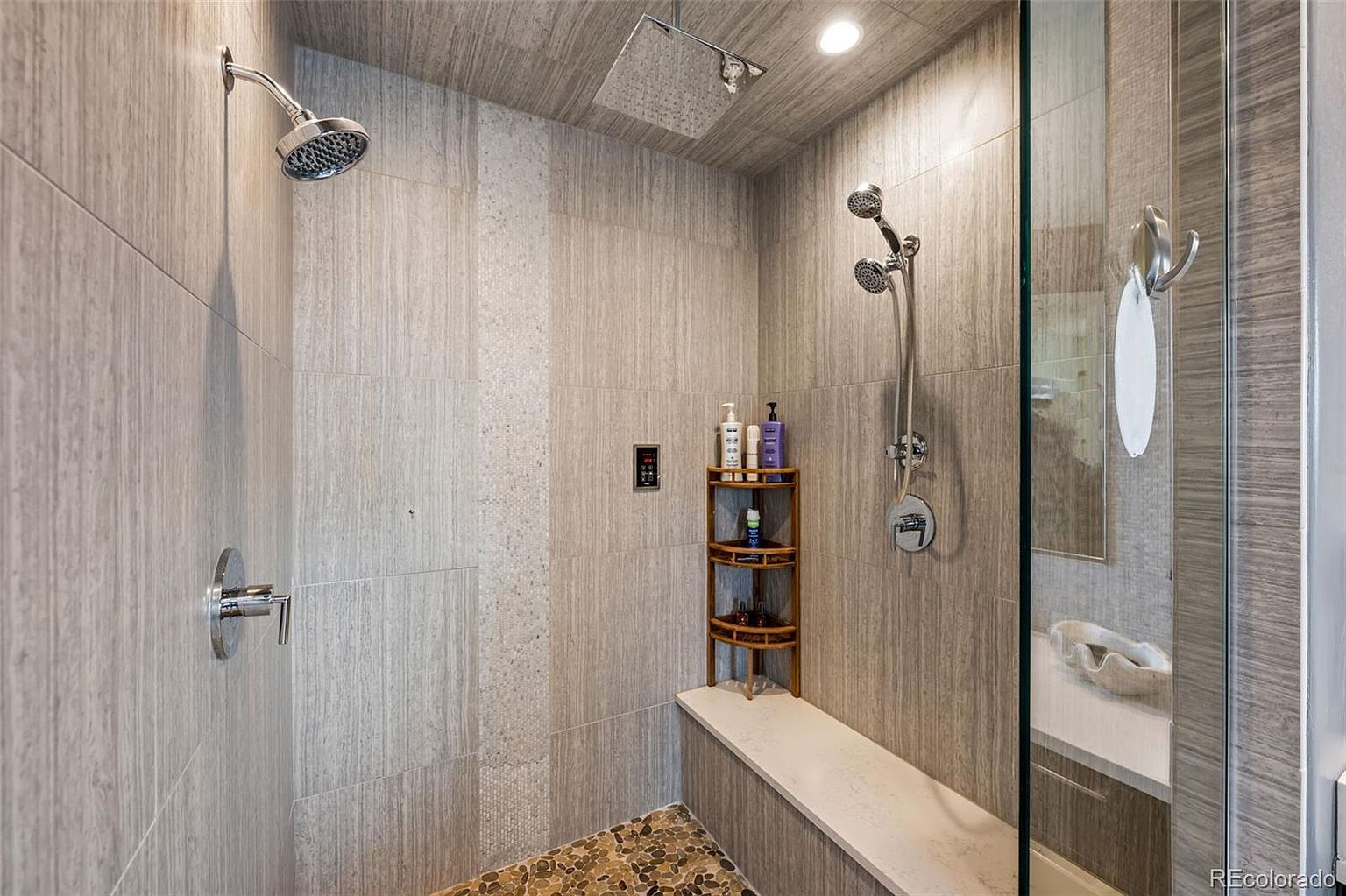 This luxurious walk-in shower features a sophisticated blend of wood-look tile walls and a natural pebble stone floor. It is equipped with multiple shower heads, including a rainfall fixture and a handheld sprayer, alongside a built-in quartz bench and a tiered wooden corner shelf. The space is illuminated by recessed lighting, creating a spa-like atmosphere with a clean, modern aesthetic.