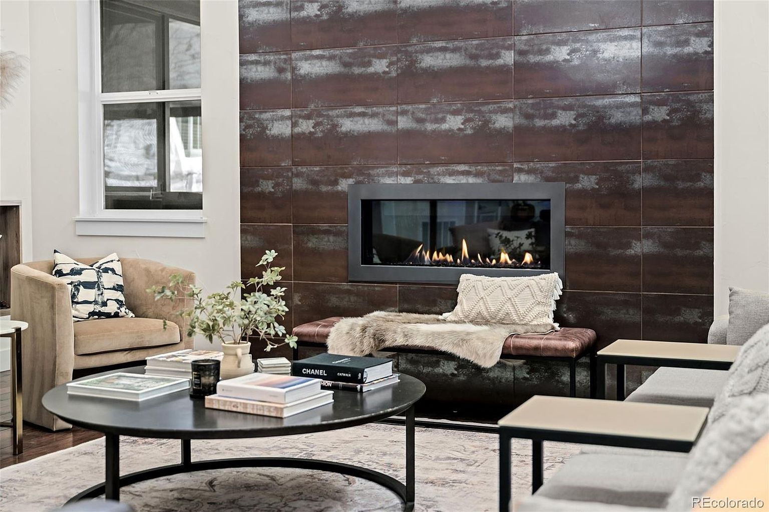 This sophisticated living room features a striking floor-to-ceiling fireplace wall clad in large, metallic-finish tiles that create a dramatic focal point. A sleek, linear gas fireplace is centered within the wall, complemented by a plush leather bench, a round coffee table adorned with coffee table books, and comfortable seating. The space exudes a modern, luxurious aesthetic with a neutral color palette and high-end textures.