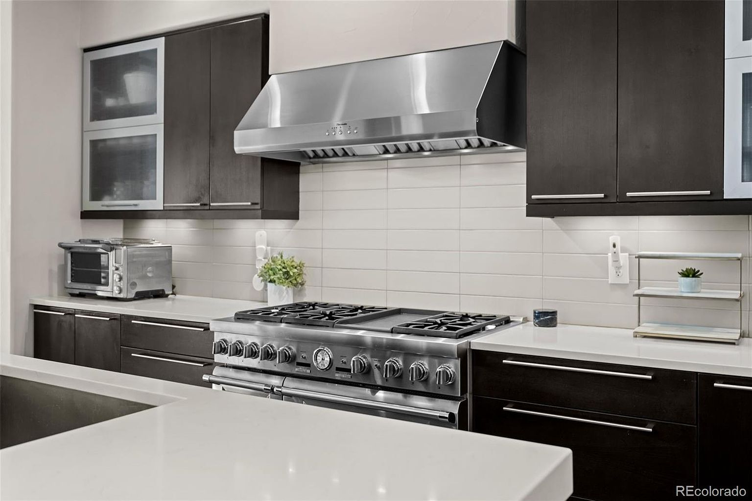 This modern kitchen features sleek, dark espresso cabinetry contrasted by bright white quartz countertops and a clean, horizontal subway tile backsplash. A professional-grade stainless steel range and matching range hood serve as the focal point, complemented by a stainless steel toaster oven and minimalist decor. The perspective is a eye-level, wide-angle shot that emphasizes the clean lines and high-end finishes of this contemporary culinary space.