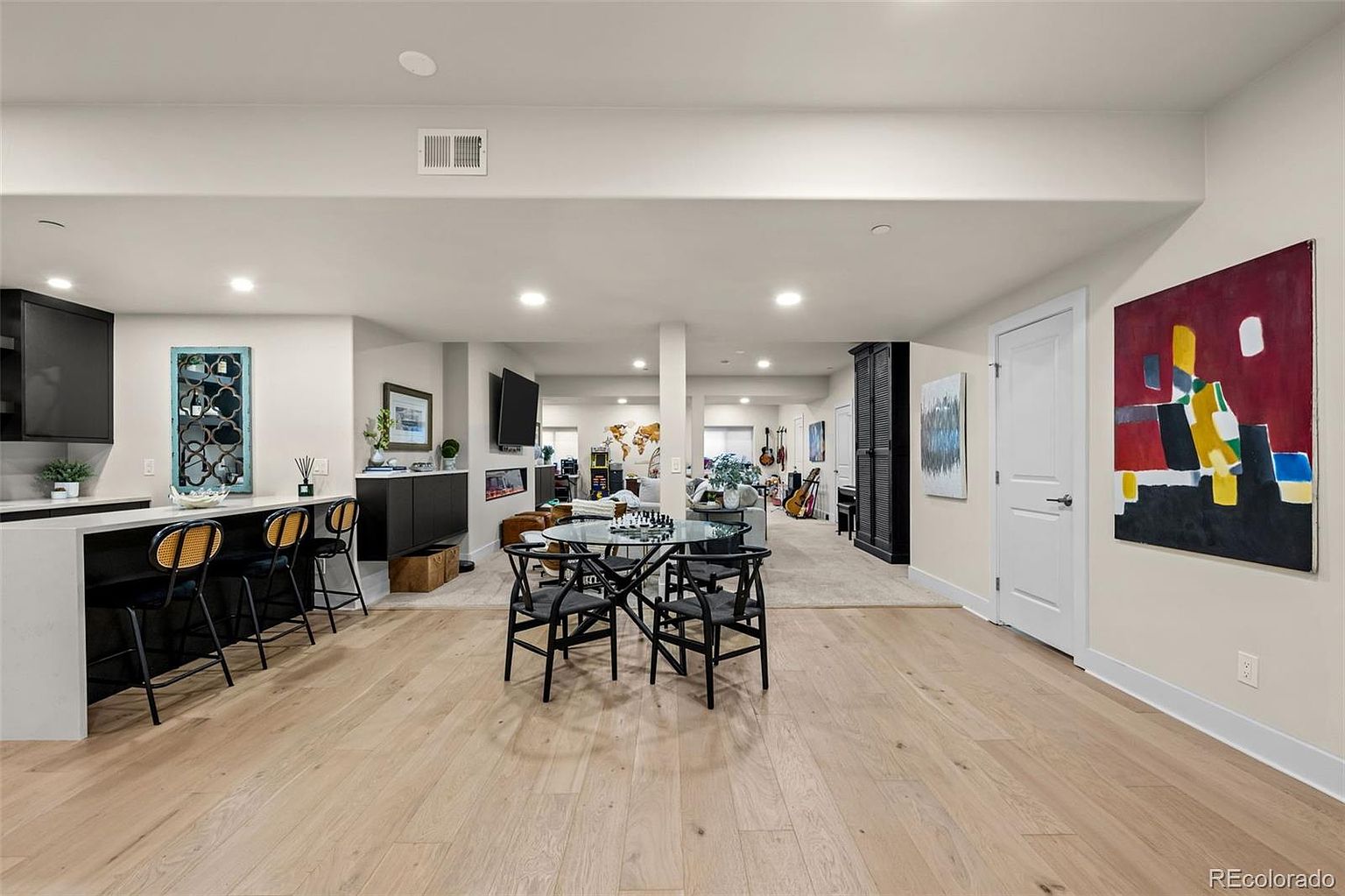 This expansive, open-concept basement features a modern aesthetic with light wood flooring and a neutral color palette. The space includes a sleek kitchen area with a breakfast bar, a central dining zone with a glass-topped table, and a comfortable living area in the background. The room is accented by vibrant abstract art and recessed lighting, creating a versatile and inviting multi-purpose living space.