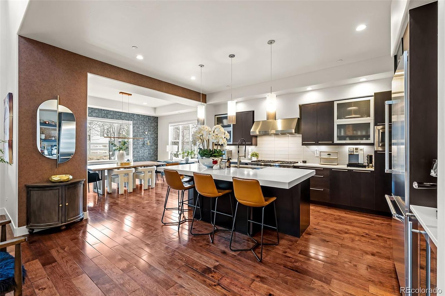 This sophisticated, open-concept kitchen features dark cabinetry contrasted by a large, white-topped central island with three modern, amber-colored leather bar stools. The space is illuminated by sleek pendant lighting and recessed ceiling lights, highlighting the rich hardwood flooring that flows seamlessly into the adjacent dining area. The design is contemporary and elegant, offering a perfect blend of functionality and high-end aesthetic appeal.
