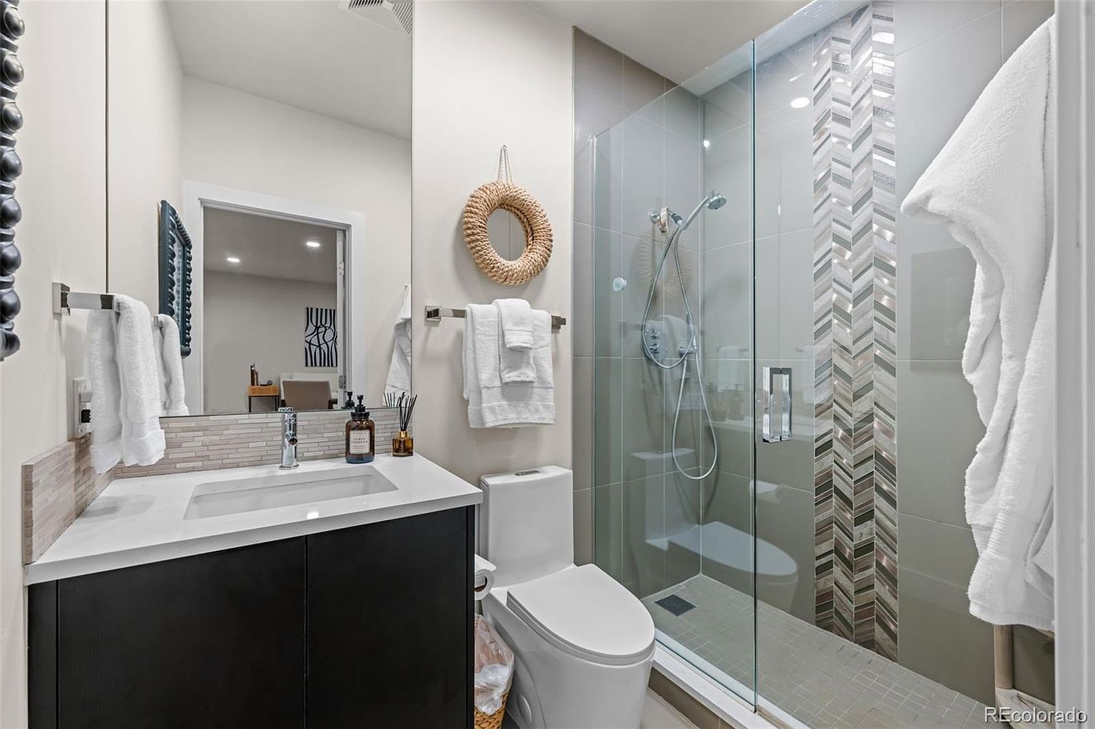 This modern bathroom features a sleek vanity with a dark cabinet, a white countertop, and a large mirror reflecting the space. To the right, a glass-enclosed walk-in shower showcases a stylish herringbone-patterned tile accent wall and a rainfall showerhead. The room is accented with a decorative woven wall hanging and plush white towels, creating a clean and contemporary aesthetic.