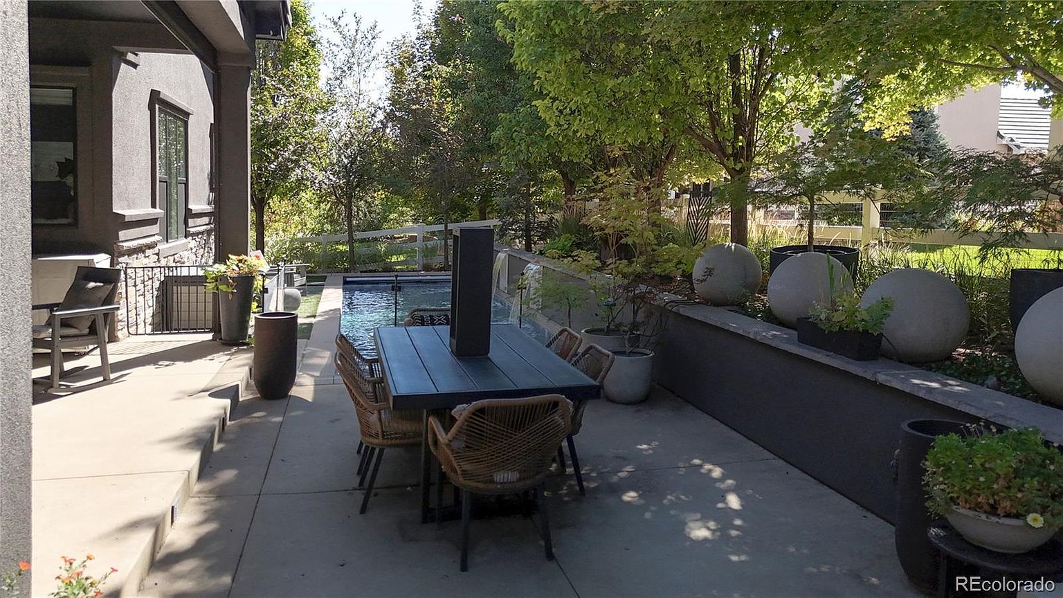 This inviting outdoor patio space features a modern dining set positioned alongside a sleek, rectangular swimming pool with a water feature. The area is framed by a low stone retaining wall adorned with decorative concrete spheres and lush greenery, creating a sophisticated and tranquil atmosphere. The perspective captures the seamless transition between the covered patio and the landscaped backyard, highlighting an ideal setting for outdoor entertaining.