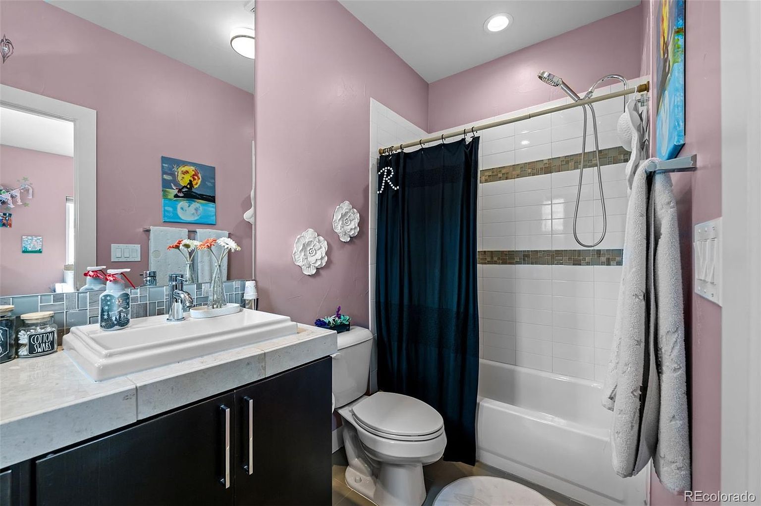 This bathroom features a modern vessel sink set atop a dark vanity with a stone countertop, complemented by soft pink walls and decorative floral wall accents. A shower-tub combination with a dark navy curtain and a handheld showerhead provides a functional and stylish bathing area. The space is well-lit with recessed lighting and includes a large mirror that enhances the overall brightness of the room.