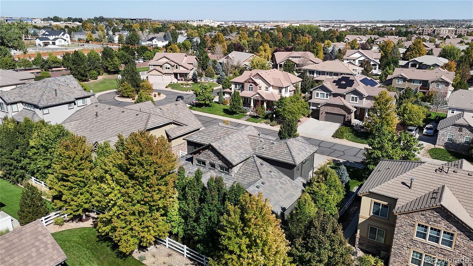 This high-angle aerial view captures a suburban neighborhood characterized by large, multi-story single-family homes with complex, multi-gabled rooflines. The scene features a mix of stone and siding exteriors, well-manicured lawns, and mature trees that provide a sense of established community. The perspective highlights the layout of the residential streets and the density of the housing development under a clear blue sky.