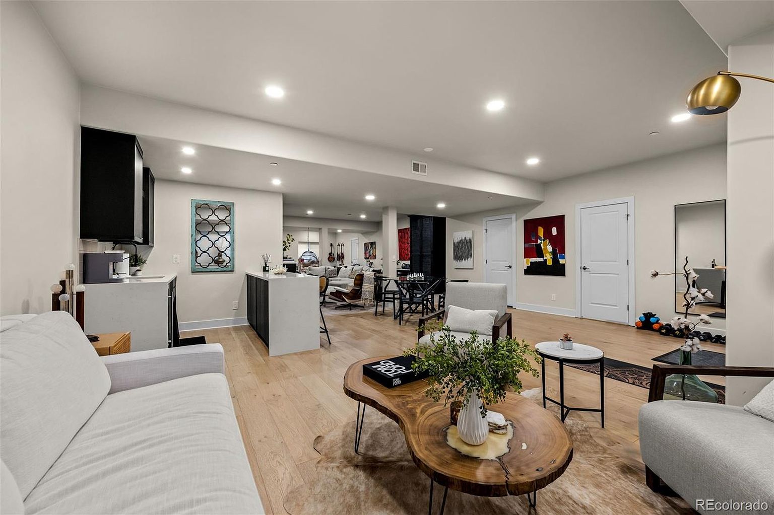 This spacious, open-concept living area features light hardwood flooring and a neutral color palette, creating a bright and airy atmosphere. The room includes a comfortable seating area with a unique live-edge wood coffee table, a wet bar with dark cabinetry, and a seamless transition into a dining space in the background. The perspective is from the foreground, looking across the living room toward the kitchen and dining area, highlighting the home's versatile and modern layout.