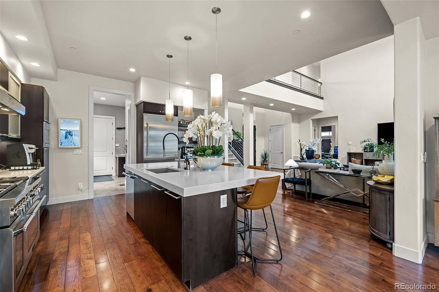 This modern, open-concept kitchen features a large central island with a white quartz countertop and dark cabinetry, serving as the focal point of the space. The kitchen flows seamlessly into a bright, high-ceilinged living area, creating an airy and inviting atmosphere perfect for entertaining. Rich hardwood flooring runs throughout, complemented by sleek pendant lighting and high-end stainless steel appliances.