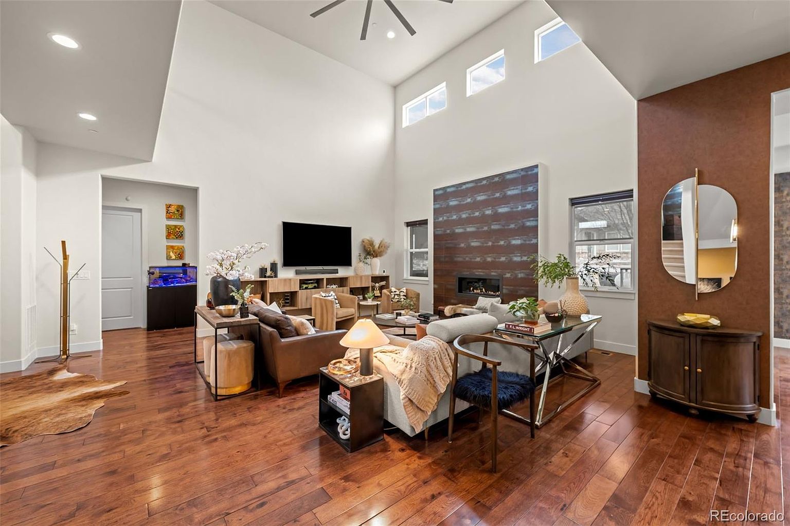 This spacious, high-ceilinged living room features warm hardwood flooring and a striking floor-to-ceiling fireplace accent wall with horizontal wood paneling. The room is furnished with a comfortable sectional sofa, a console table, and a media center, all bathed in natural light from large clerestory windows. A decorative mirror and a curved cabinet add a touch of elegance to the side wall, creating a welcoming and sophisticated atmosphere.