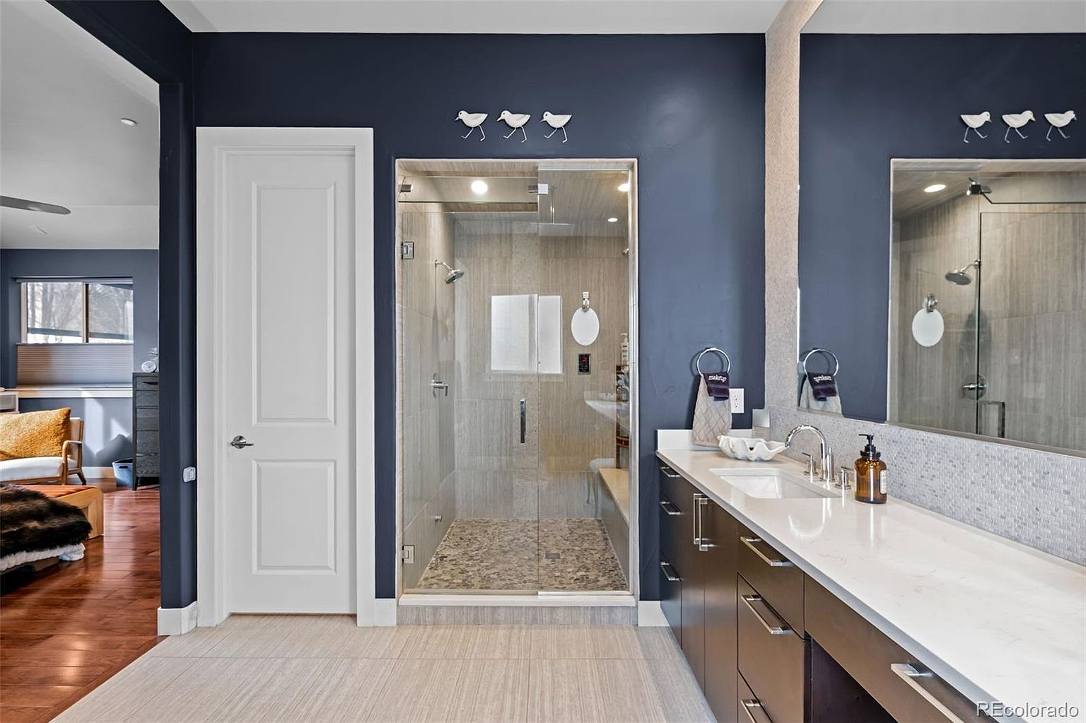 This modern primary bathroom features a sleek, dark navy accent wall contrasted by a white door and a long vanity with a white quartz countertop. A glass-enclosed walk-in shower with pebble flooring and neutral tile work serves as the focal point, while a large mirror reflects the space, enhancing the open feel. The room is well-lit and stylish, offering a clean, contemporary aesthetic.