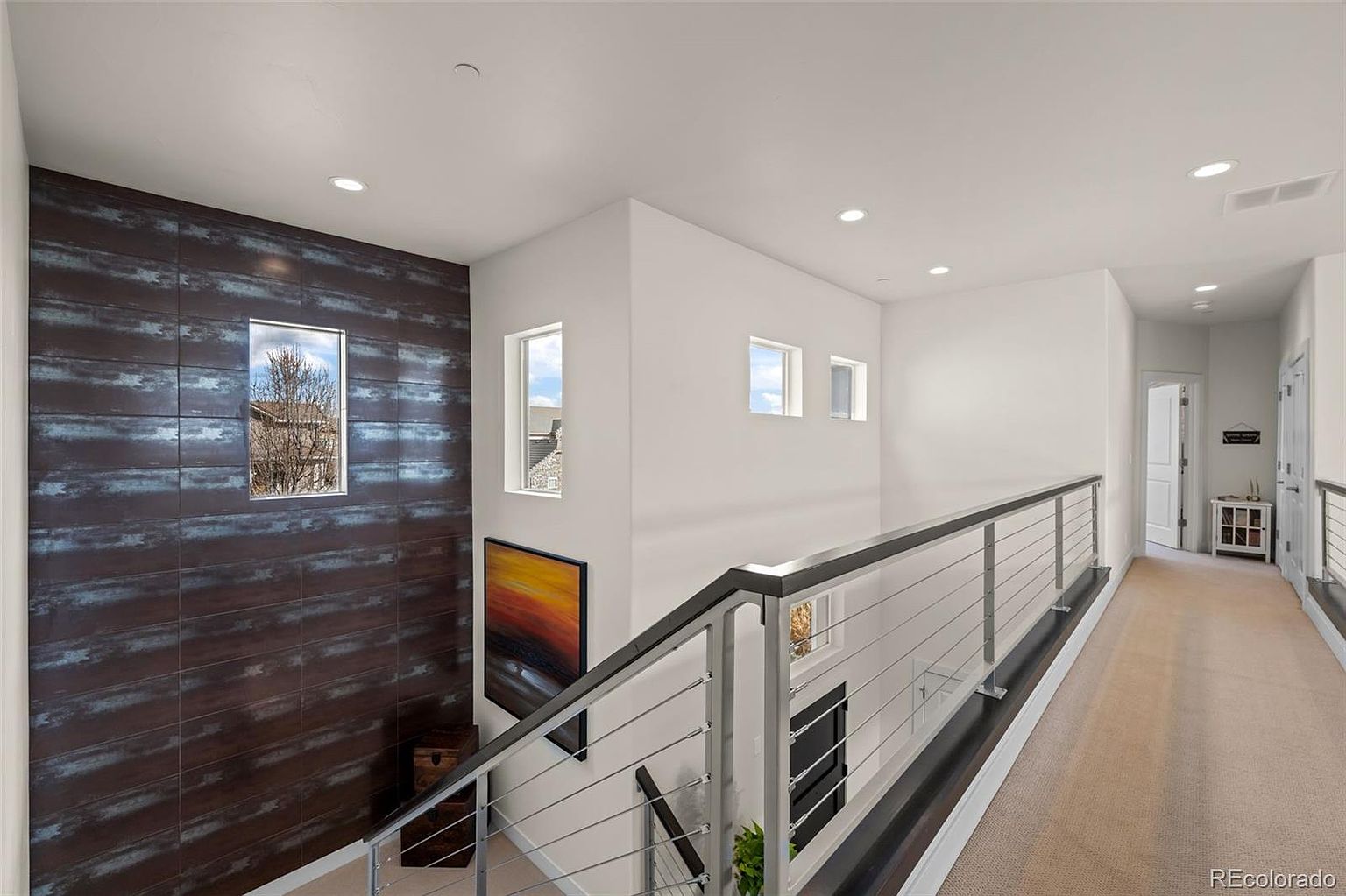 This modern hallway overlooks a staircase, featuring a striking accent wall with dark, textured horizontal tiles and a large window that brings in natural light. The space is defined by a sleek metal cable railing, neutral carpeting, and recessed lighting, creating a clean and contemporary aesthetic. The perspective captures the transition between levels, highlighting the architectural openness and sophisticated design of the home.
