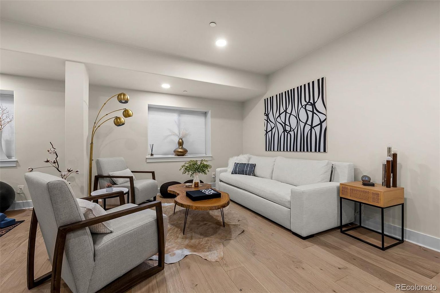 This bright and inviting living room features a contemporary design with light wood flooring, a neutral color palette, and a comfortable white sofa. The space is accented by a unique abstract black-and-white wall art piece, a natural wood coffee table, and a stylish gold floor lamp. The perspective is a wide-angle shot that captures the cozy seating arrangement, making the room feel open and well-curated for relaxation.