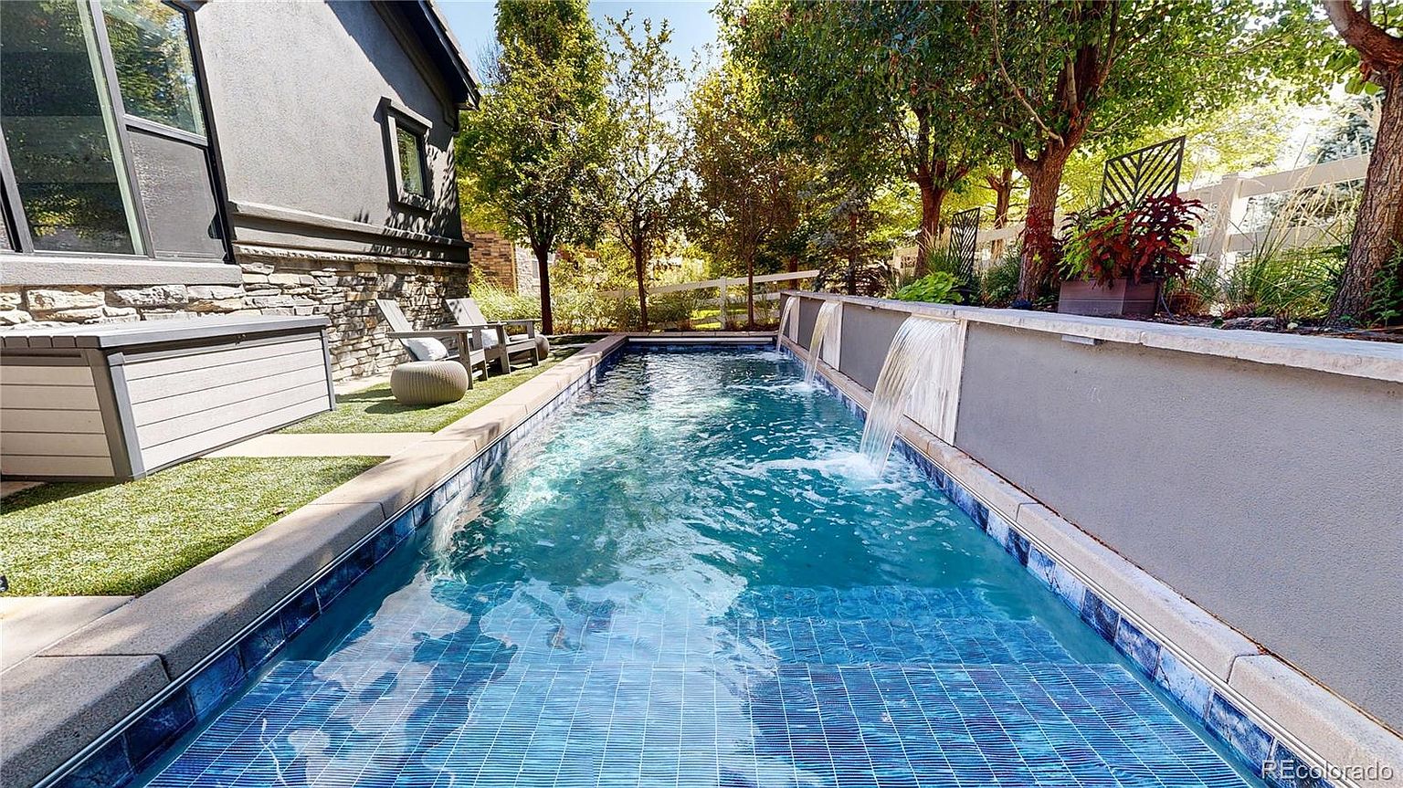 This image showcases a sleek, modern lap pool featuring vibrant blue mosaic tiles and integrated water features that cascade from a raised stone wall. The pool area is complemented by a narrow strip of artificial turf, a storage bench, and comfortable outdoor seating, all set against a backdrop of lush trees and a white fence. The perspective is low and centered, emphasizing the length of the pool and the tranquil, resort-like atmosphere of the backyard.