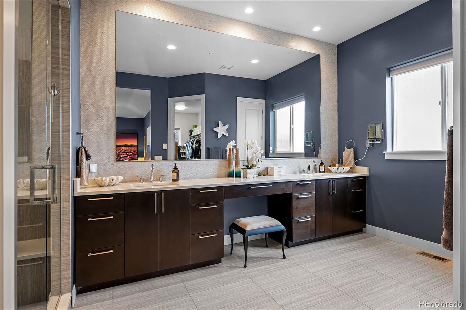 This spacious primary bathroom features a long, dark wood vanity with a white countertop, dual sinks, and a central makeup station. A large, frameless mirror spans the wall above the vanity, reflecting the deep blue walls and a bright, airy window. The room is finished with light-toned floor tiles and a glass-enclosed shower, creating a clean, modern, and sophisticated aesthetic.