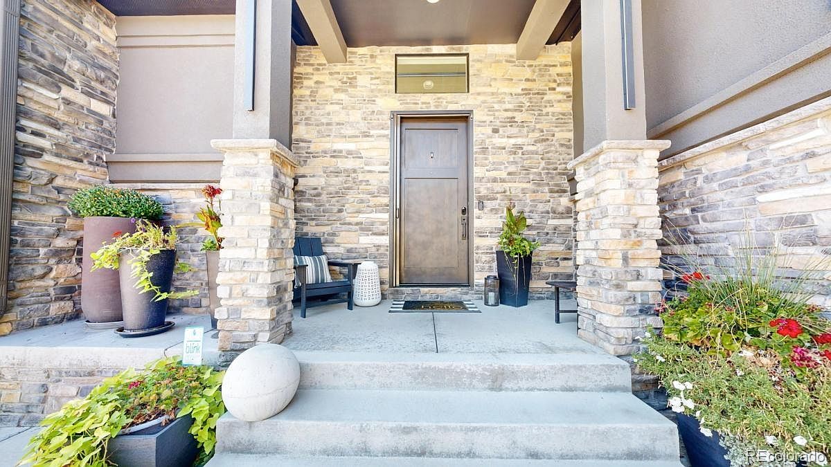 This inviting front entryway features a sturdy, dark wood door framed by rustic stone veneer siding and substantial stone-clad pillars. The covered porch includes a comfortable seating area with a dark chair and potted plants, creating a warm and welcoming atmosphere. The perspective is a straight-on, eye-level shot that highlights the architectural textures and the clean, modern aesthetic of the home's entrance.