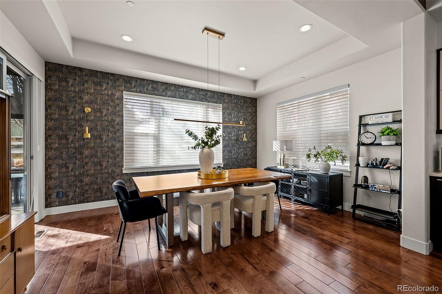 This elegant dining room features a warm, natural wood table paired with modern, cream-colored upholstered chairs, set against a striking dark textured accent wall. The space is illuminated by a sleek, minimalist linear pendant light and complemented by a black metal shelving unit and a matching sideboard. The rich hardwood flooring and abundant natural light from the large windows create a sophisticated and inviting atmosphere.