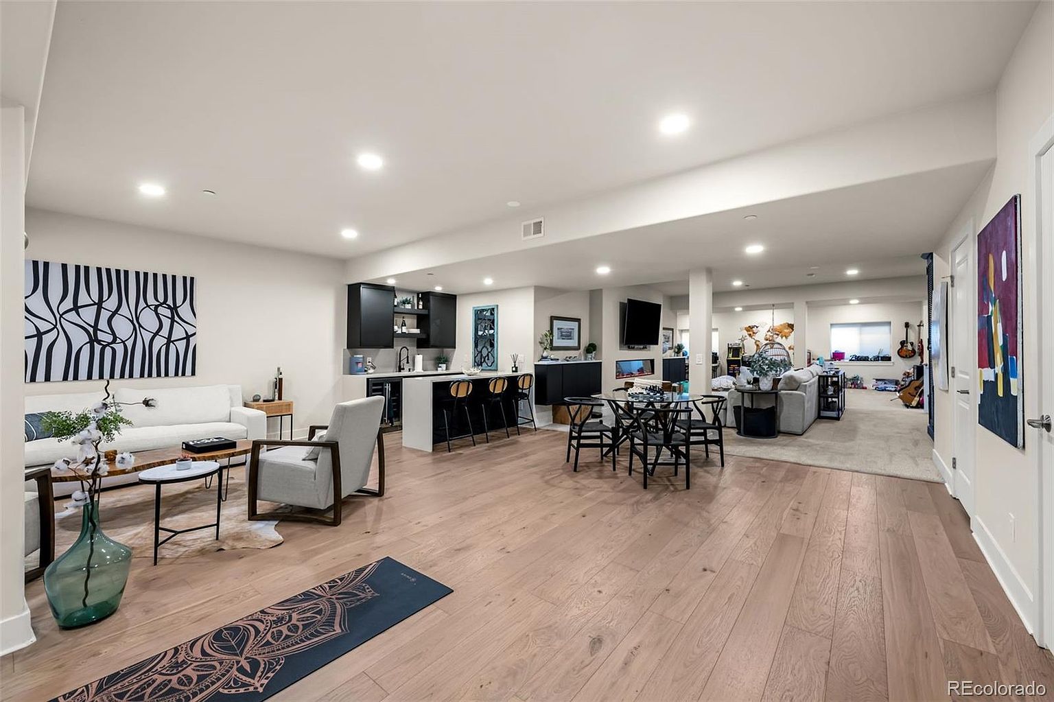 This expansive, open-concept basement features a versatile layout that seamlessly integrates a cozy living area, a sleek wet bar, and a dining space. The room is finished with light wood flooring, recessed lighting, and contemporary decor, creating a bright and inviting atmosphere perfect for entertaining. The perspective captures the entire floor plan, showcasing the transition from the lounge area to the secondary living space in the background.