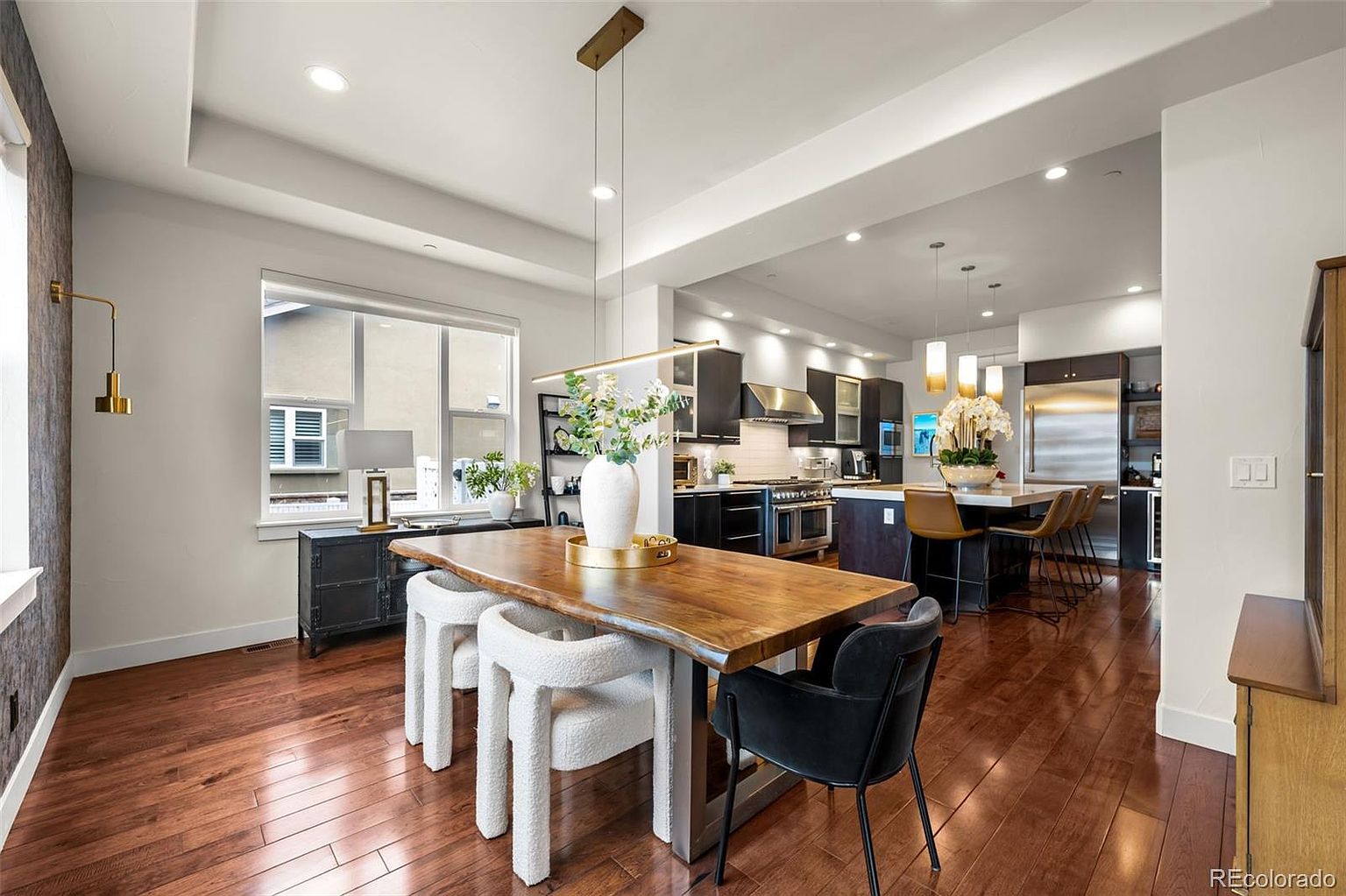 This open-concept dining area features a rustic live-edge wood table paired with modern, textured white chairs and a sleek black armchair. The space flows seamlessly into a contemporary kitchen with dark cabinetry, stainless steel appliances, and a large island with bar seating. Warm hardwood flooring and sophisticated pendant lighting create an inviting, high-end atmosphere perfect for entertaining.