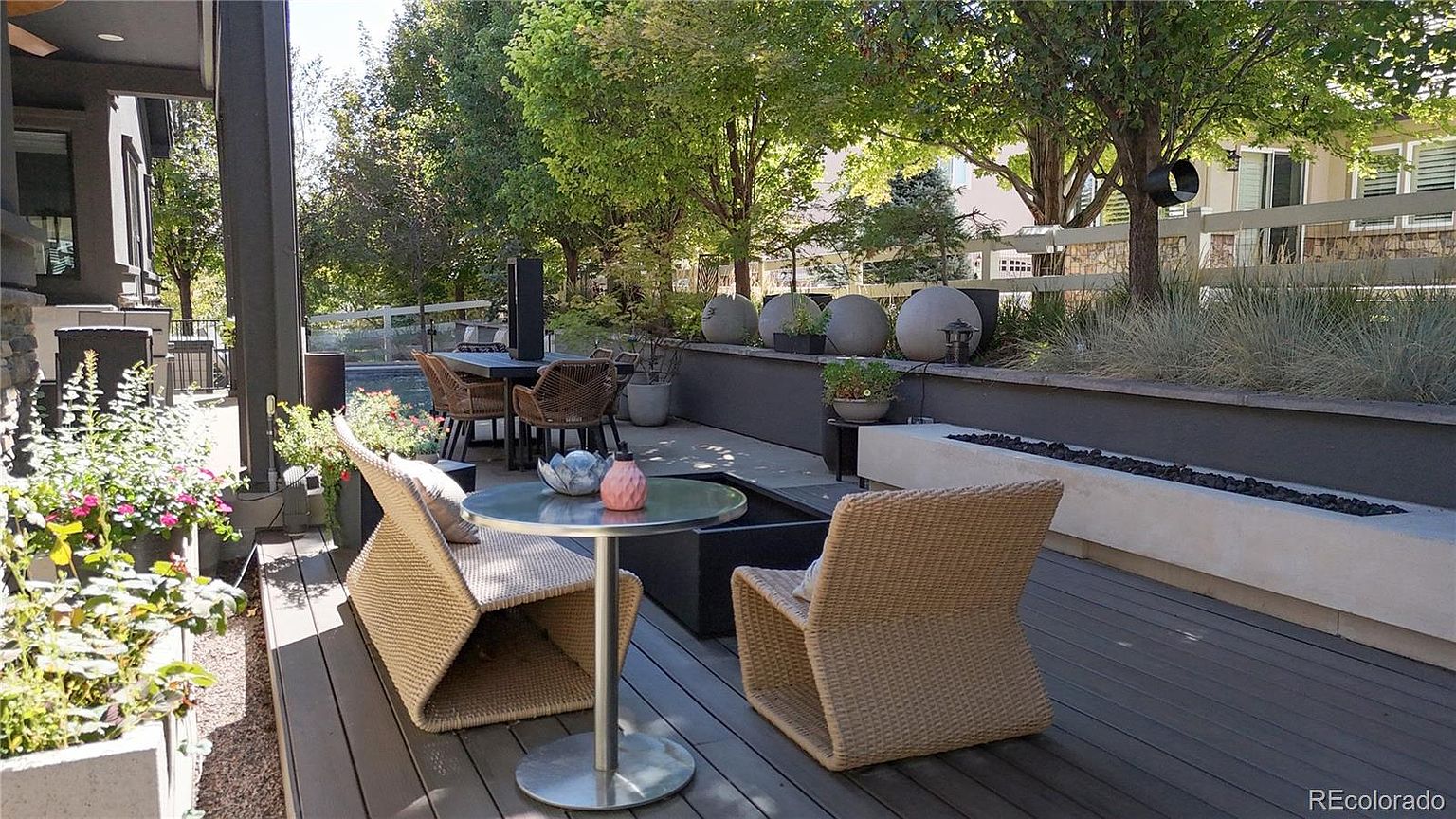 This inviting outdoor living space features a modern, multi-level deck with a comfortable seating area centered around a sleek fire pit. The area is accented by lush greenery, decorative stone spheres, and a dining set in the background, creating a sophisticated and tranquil atmosphere perfect for entertaining. The perspective captures the seamless transition between the indoor living area and the well-manicured backyard.