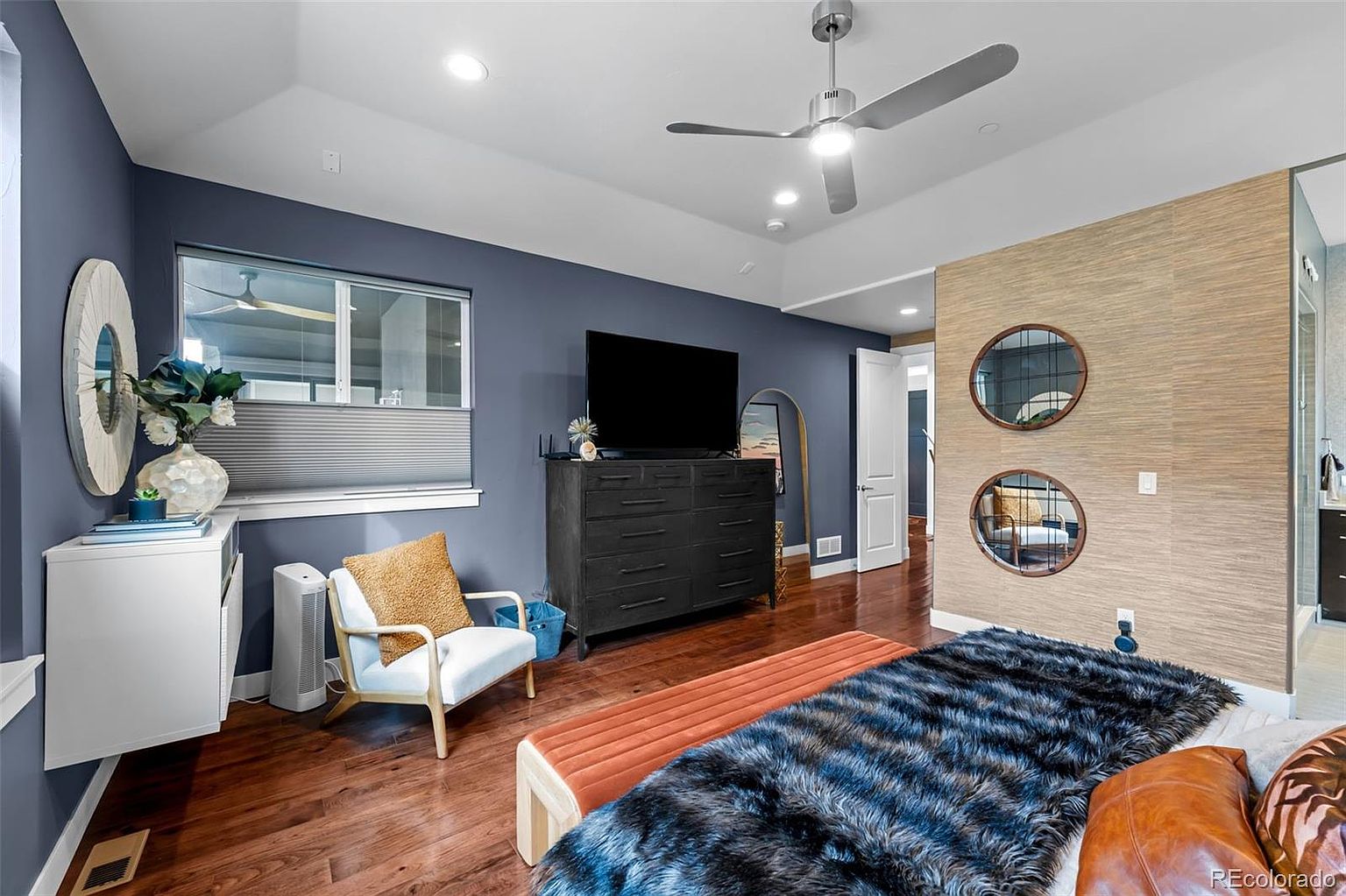 This spacious primary bedroom features a sophisticated design with a deep blue accent wall, rich hardwood flooring, and a textured wood-paneled wall behind the bed. The room is furnished with a modern black dresser, a cozy armchair, and a plush bed with a faux fur throw, all illuminated by a contemporary ceiling fan. The perspective captures the inviting layout, highlighting the blend of modern textures and warm, comfortable aesthetics.