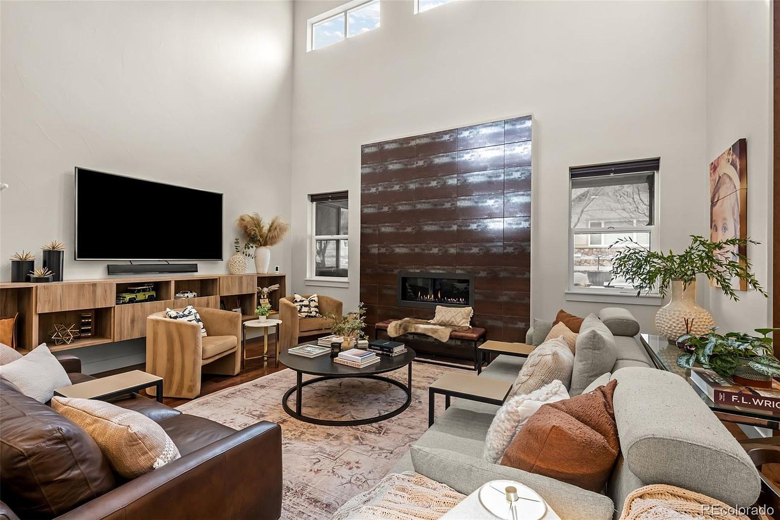 This spacious, high-ceilinged living room features a striking floor-to-ceiling dark metal fireplace surround that serves as a dramatic focal point. The room is furnished with a comfortable grey sectional, a leather sofa, and two tan accent chairs arranged around a circular coffee table, creating a warm and inviting atmosphere. Large windows allow for ample natural light, while the open-concept layout and modern decor provide a sophisticated yet cozy living space.