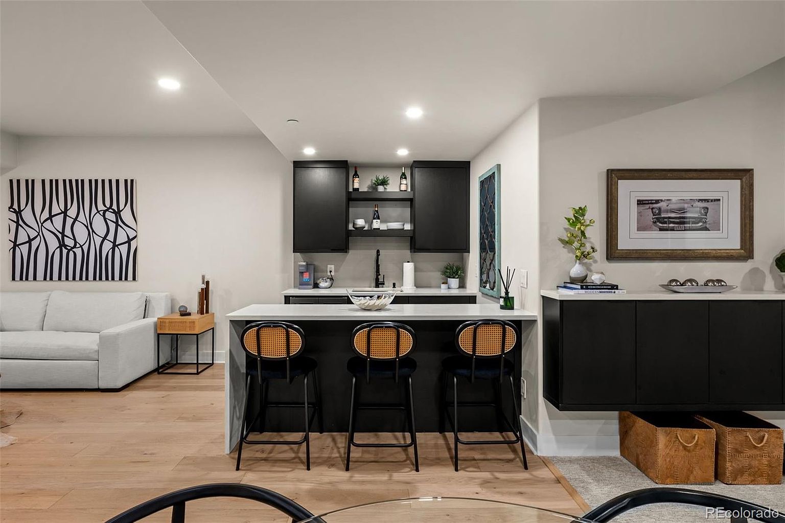 This modern basement living area features a sleek wet bar with dark cabinetry, a white quartz countertop, and three stylish cane-back bar stools. To the left, a comfortable light-colored sofa sits adjacent to a large, bold abstract art piece, while the right side showcases a framed vintage car print above a black storage console. The space is illuminated by recessed lighting, creating a sophisticated and inviting atmosphere perfect for entertaining.
