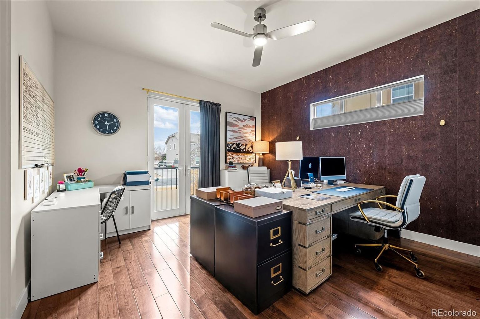This well-organized home office features a large, rustic-style wooden desk paired with a modern white and gold office chair, set against a striking dark, textured accent wall. The room includes a secondary white desk workspace, a filing cabinet, and a glass door leading to a balcony, creating a bright and productive atmosphere. The space is thoughtfully decorated with a wall clock, framed artwork, and a ceiling fan, offering a professional yet comfortable environment.