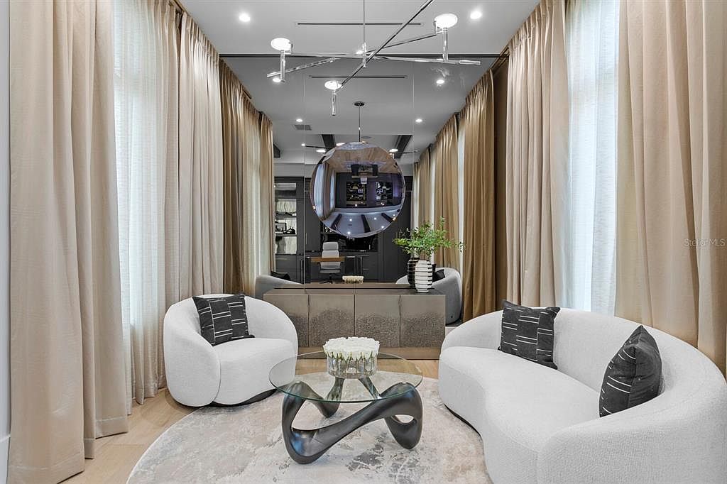 This is an interior shot of a modern living room featuring a curved white sofa and a matching armchair, both accented with patterned pillows. A unique glass coffee table sits on a round rug, and the room is framed by elegant beige curtains. A striking chandelier and a large spherical mirror add to the room's contemporary aesthetic.