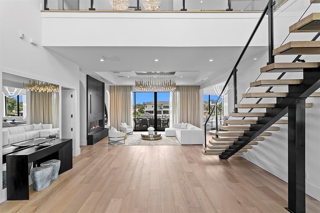 This is a spacious and luxurious living room featuring an open floor plan with hardwood floors and high ceilings. The room includes a modern fireplace, elegant chandeliers, and large windows that provide ample natural light and views of the outdoors. A contemporary staircase adds a striking architectural element to the space.