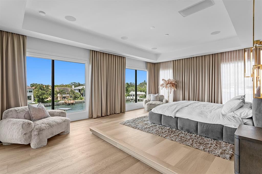 This is a luxurious primary bedroom featuring a large platform bed with a gray upholstered base and white bedding. The room has large windows with views of the water and is decorated with neutral-toned curtains and plush seating. The overall impression is one of comfort and elegance.