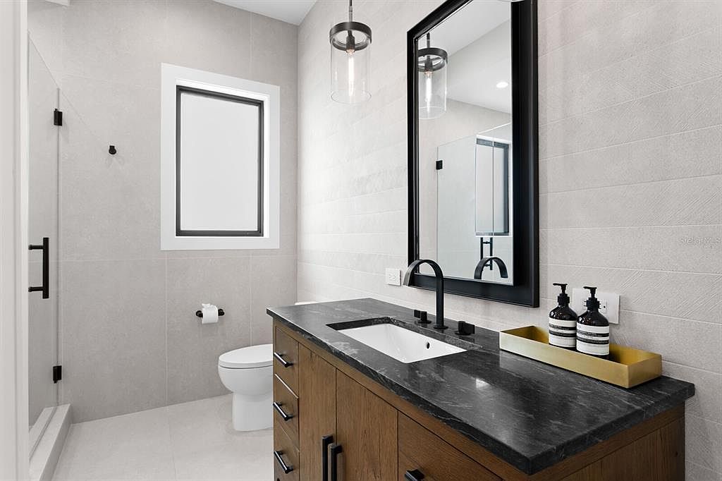 This is a modern bathroom featuring a wood vanity with a dark countertop and a white undermount sink. A black framed mirror hangs above the vanity, complemented by black fixtures and hardware throughout the space. The walls are tiled in a light gray horizontal pattern, and the room includes a toilet and a glass-enclosed shower.