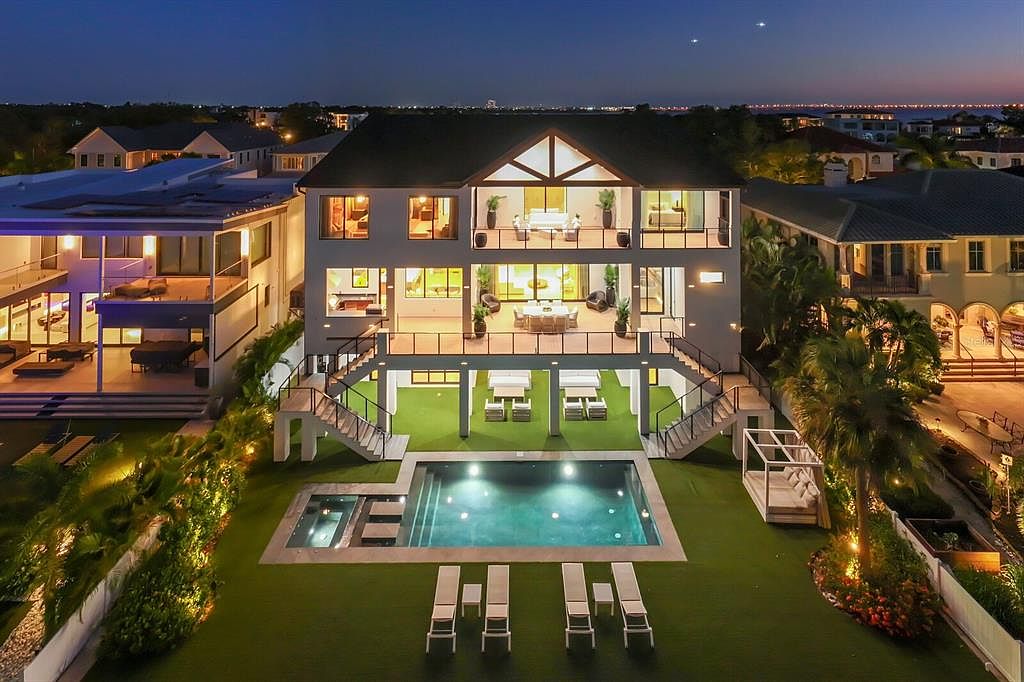 This is a rear view of a luxurious multi-story home at dusk, showcasing a modern design with large windows and multiple balconies. The property features a swimming pool, spa, and meticulously landscaped yard with lounge chairs and a covered outdoor seating area. The overall impression is one of high-end living and outdoor entertainment.